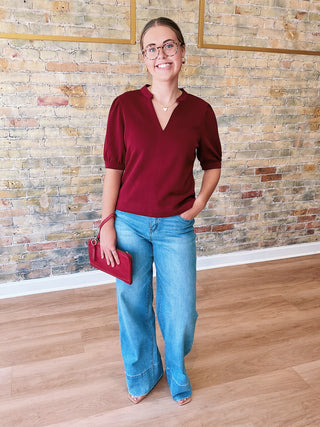 Woman in a red shirt and blue jeans standing against a brick wall.