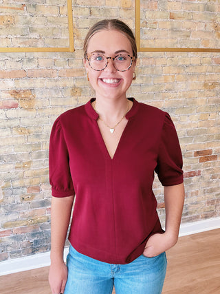 Person wearing a red blouse and blue jeans standing in front of a stone wall.