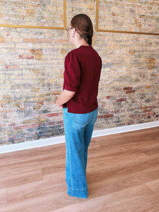Person wearing a red shirt and blue jeans standing in front of a brick wall.